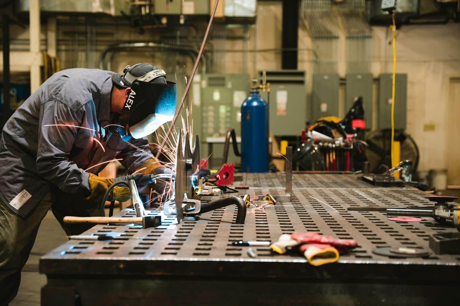 A welder in a helmet working at a steel fabrication table